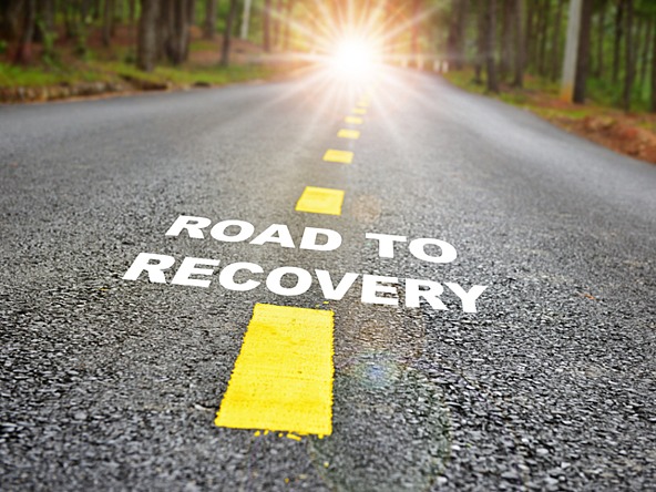 The words 'road to recovery' printed on the middle of a road, with sunshine in distance